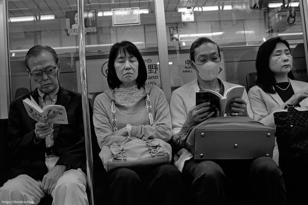 Quatre passagers âgés assis dans une rame de métro à Tokyo, certains lisant, d'autres assoupis, devant le panneau de la station Nakano.